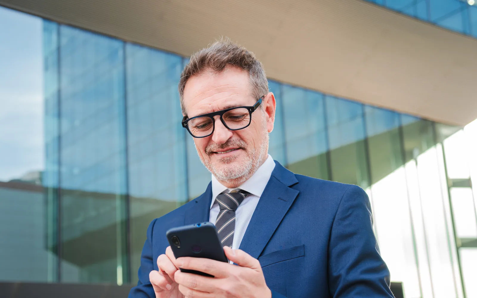 Business owner checking his company’s mobile app on a smartphone outside a glass office building