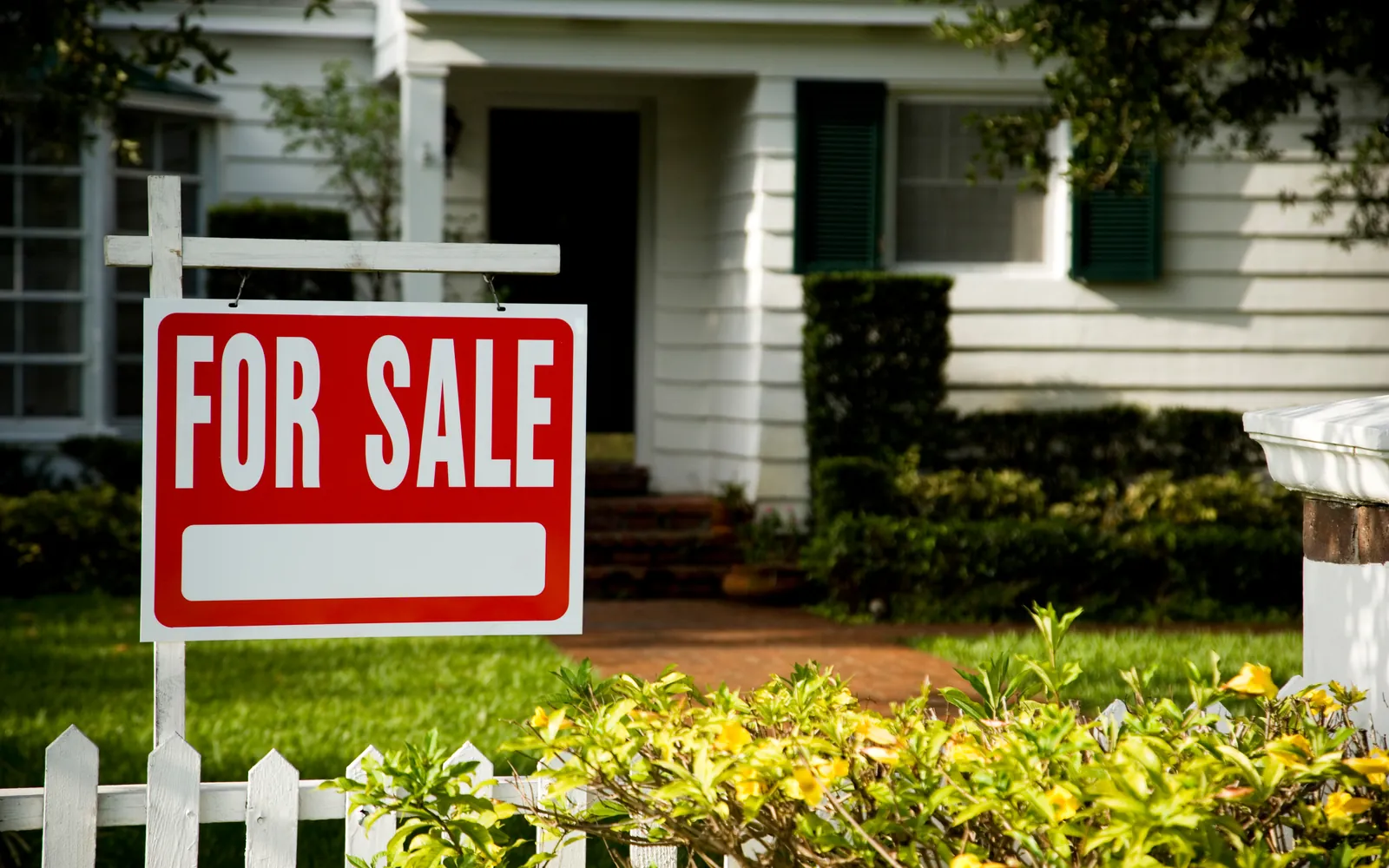 For Sale sign in front of a suburban American home with a white picket fence